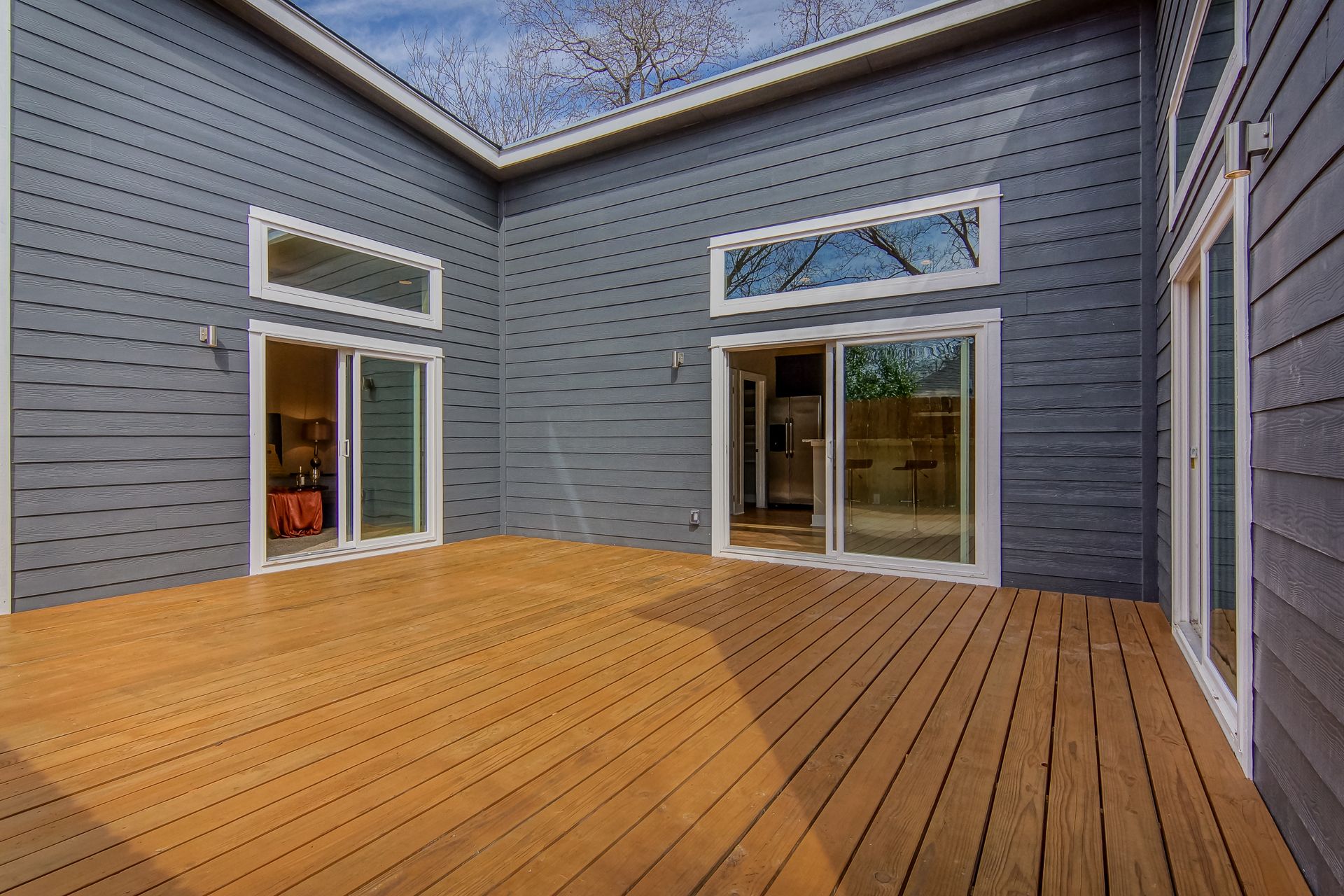 A large, rectangular wooden deck attached to a dark blue modern house with two sliding glass doors and transom windows.