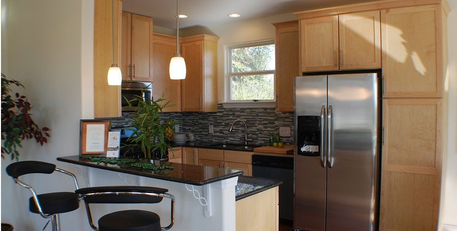 A modern kitchen with light wood cabinets, stainless steel appliances, a dark granite counter, and two bar stools.