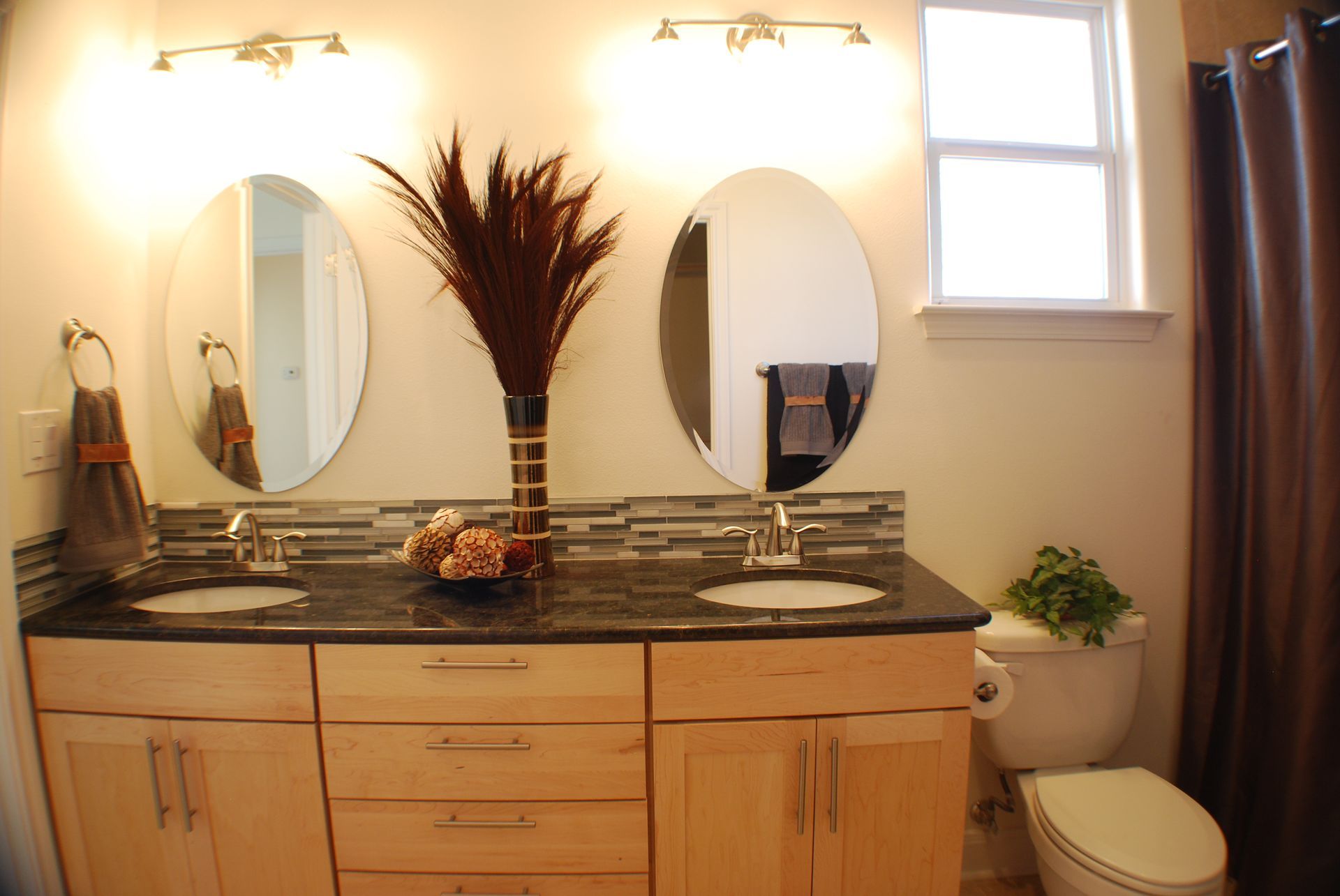 A double-vanity bathroom featuring light wood cabinets, black granite countertops, dual oval mirrors, and a white toilet.