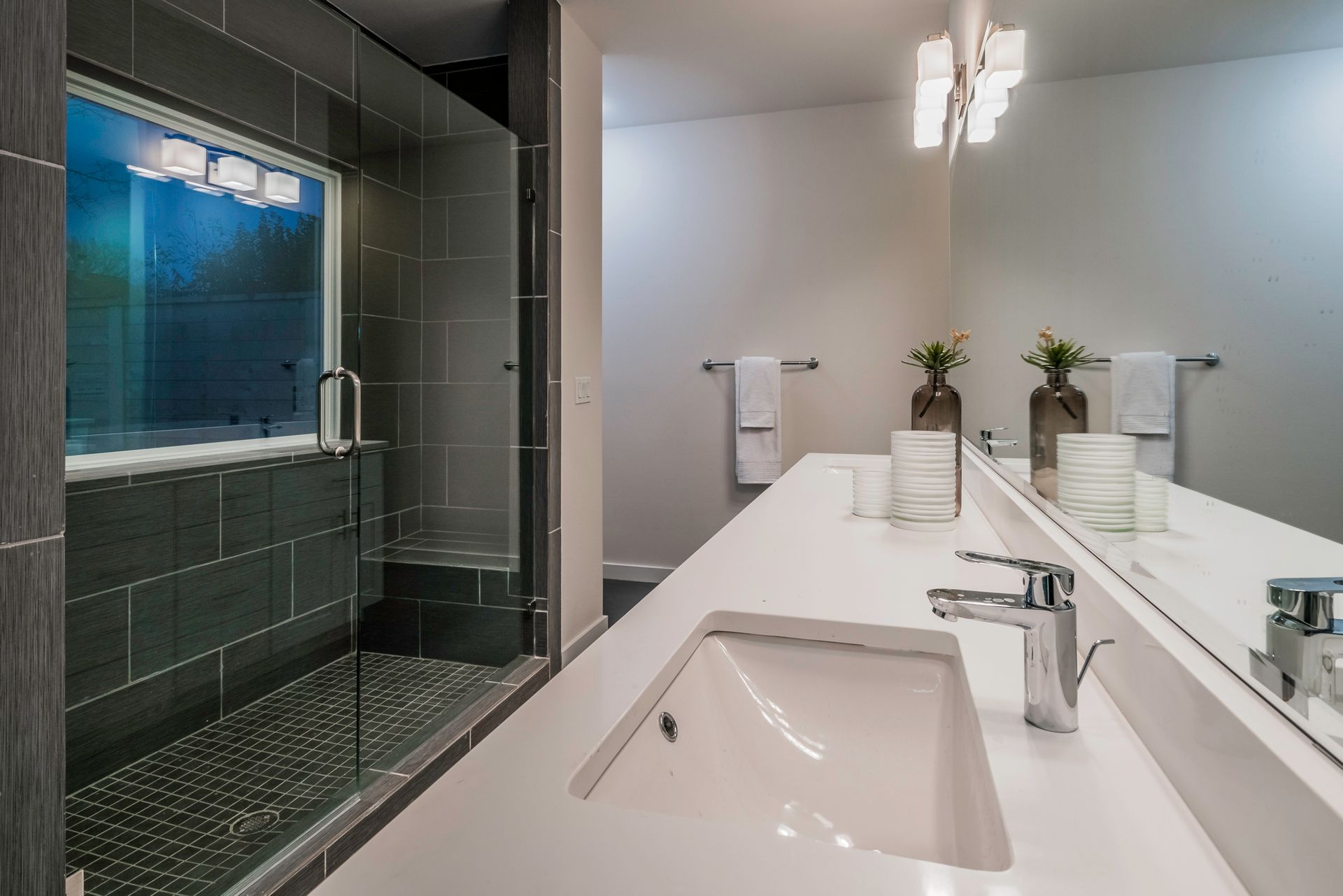Modern bathroom featuring a dark tiled walk-in shower with a glass door, white countertop, and a double vanity mirror.