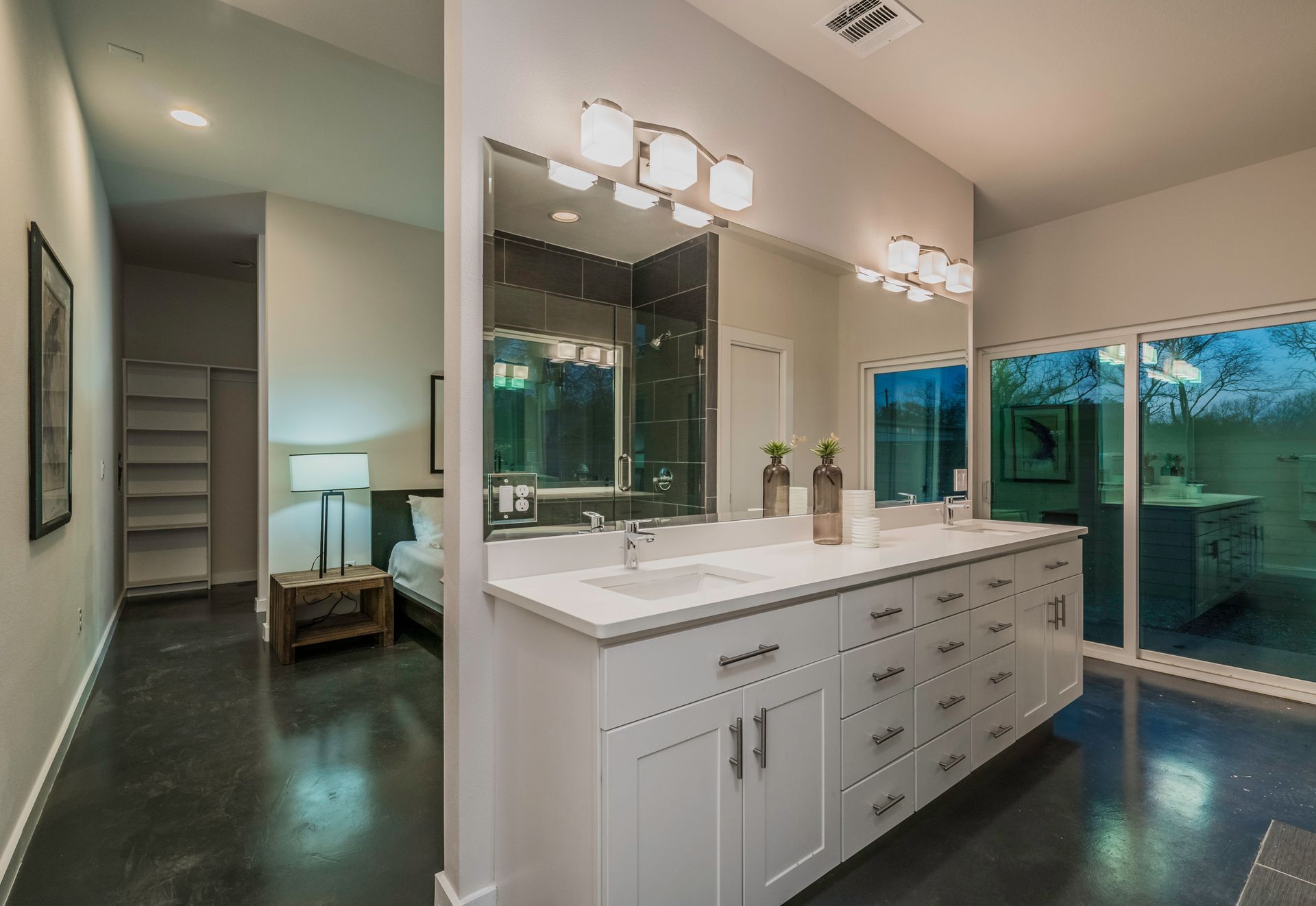 Modern bathroom with white double vanity, large mirror, and dark flooring, leading into an adjacent bedroom.