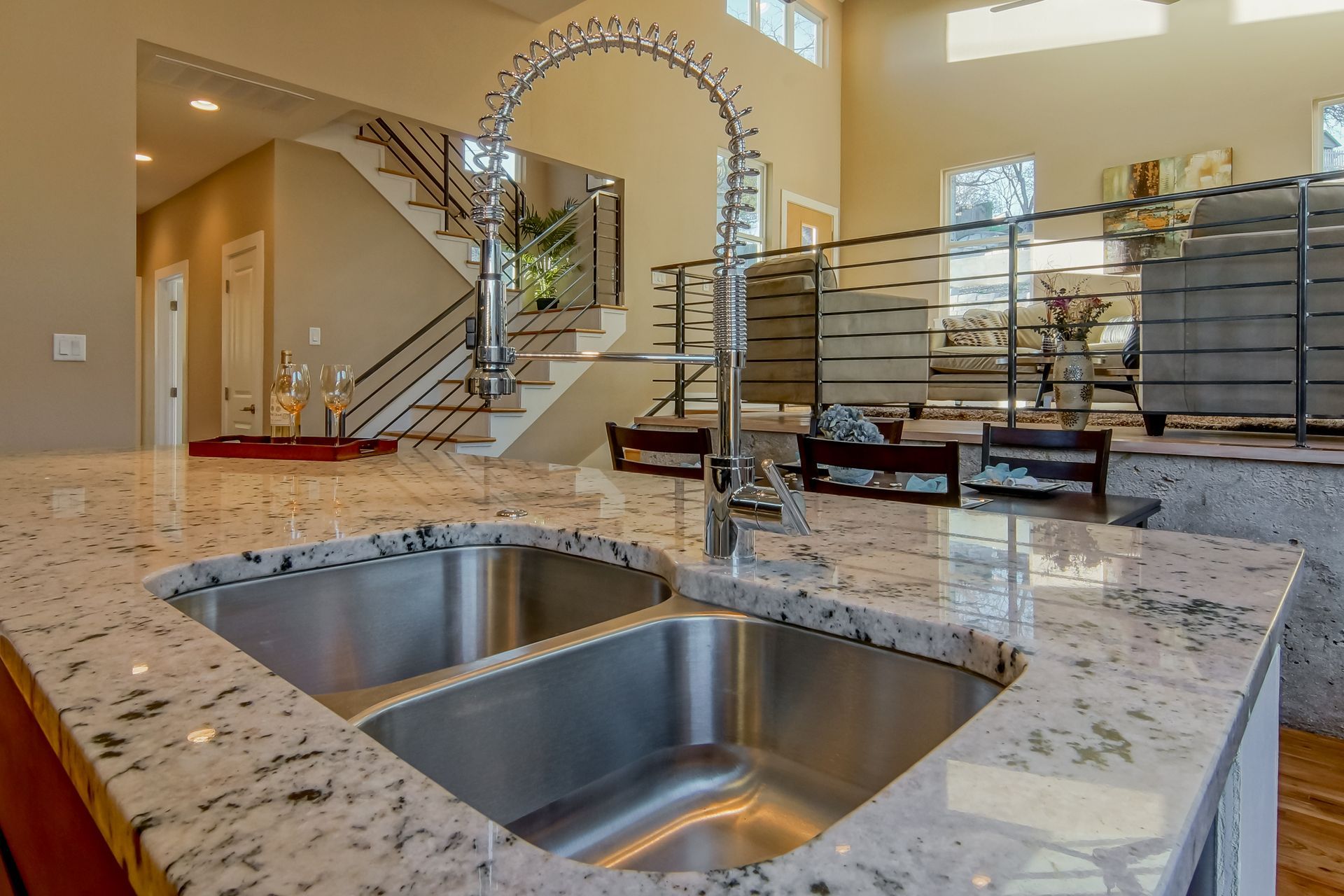 Close-up of a kitchen sink with speckled granite countertops, overlooking an open-plan living area and staircase.