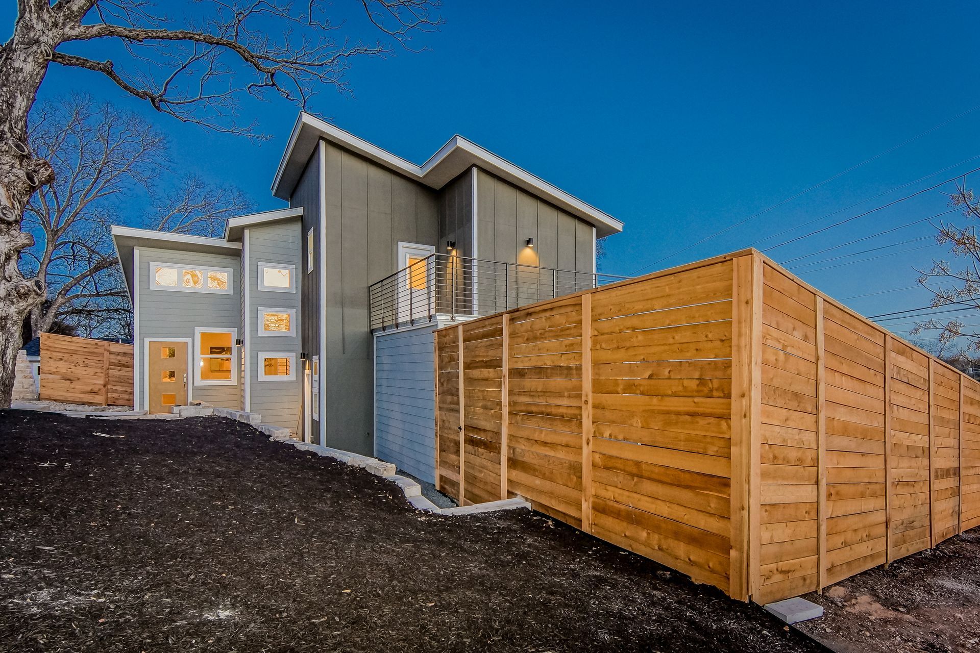 A modern, multi-story gray home with wood accents and a tall wooden privacy fence under a clear blue evening sky.