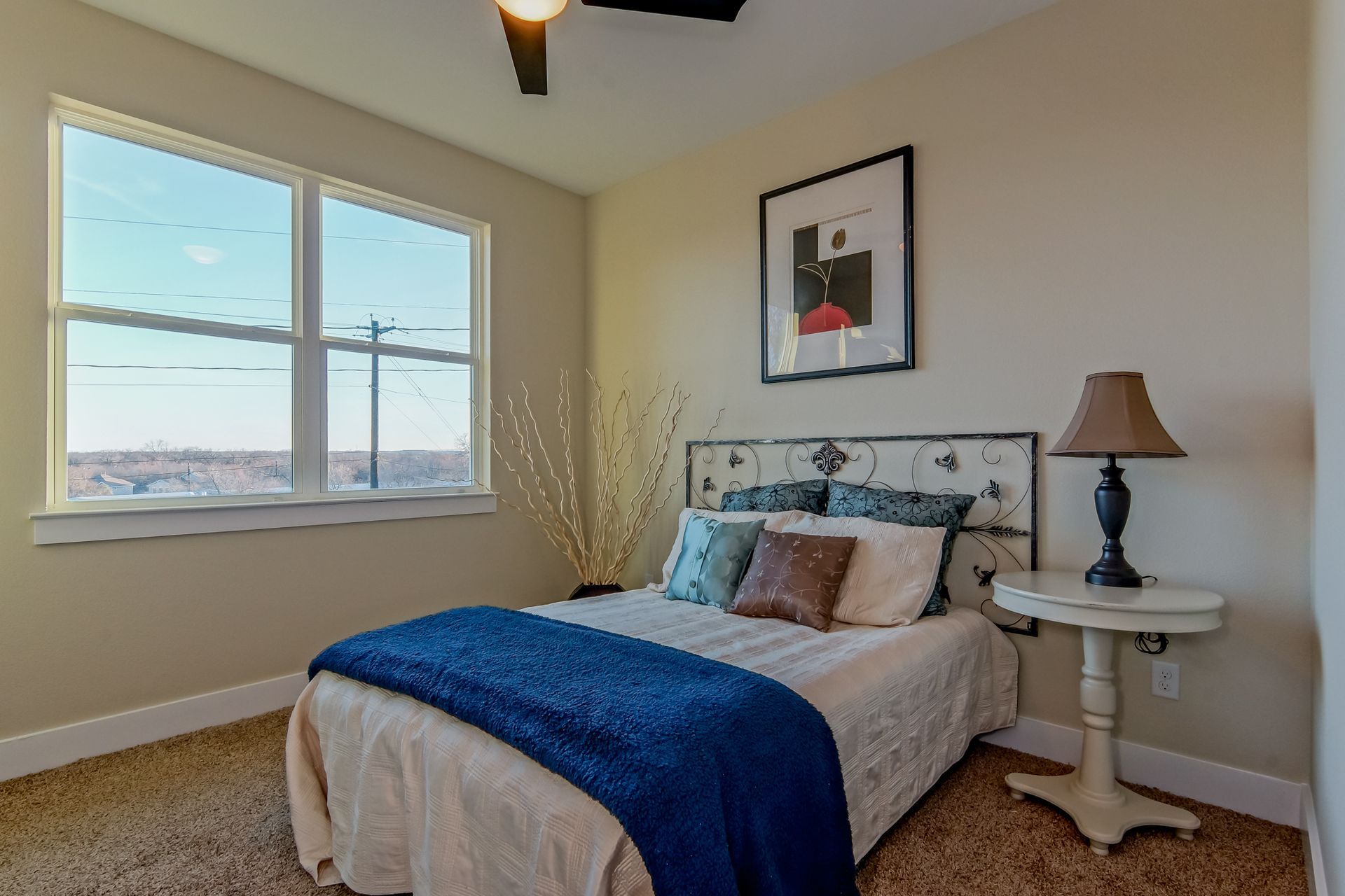A cozy bedroom with a bed topped with a blue throw blanket, a lamp on a round side table, and a window overlooking a view.