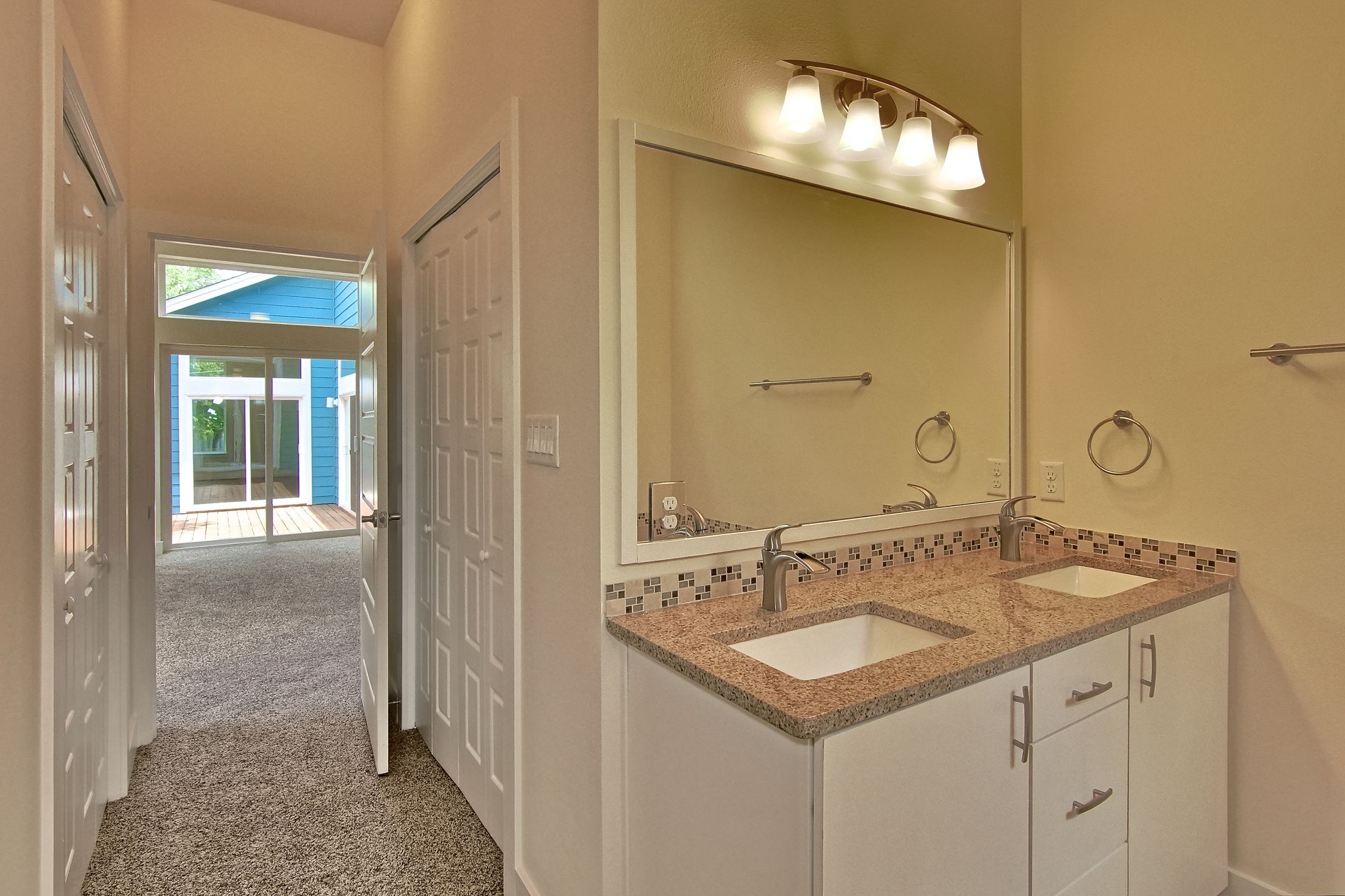 A bathroom vanity with dual sinks and a granite countertop next to a doorway looking out into a carpeted living area.