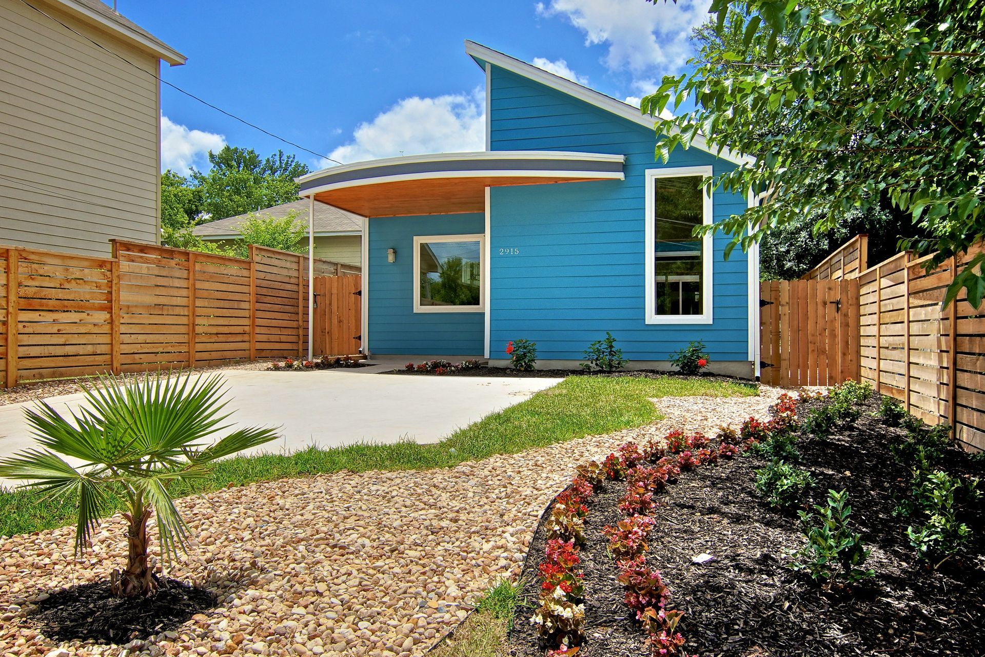 A bright blue modern house with a slanted roof and curved porch, set behind a landscaped yard with rocks and a palm tree.