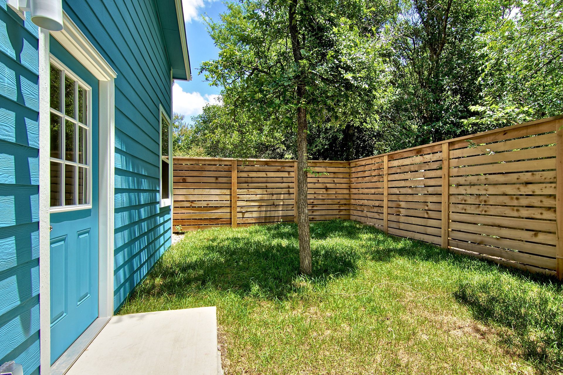 A blue house exterior with a small yard, green grass, and a wooden horizontal slat privacy fence under a sunny sky.