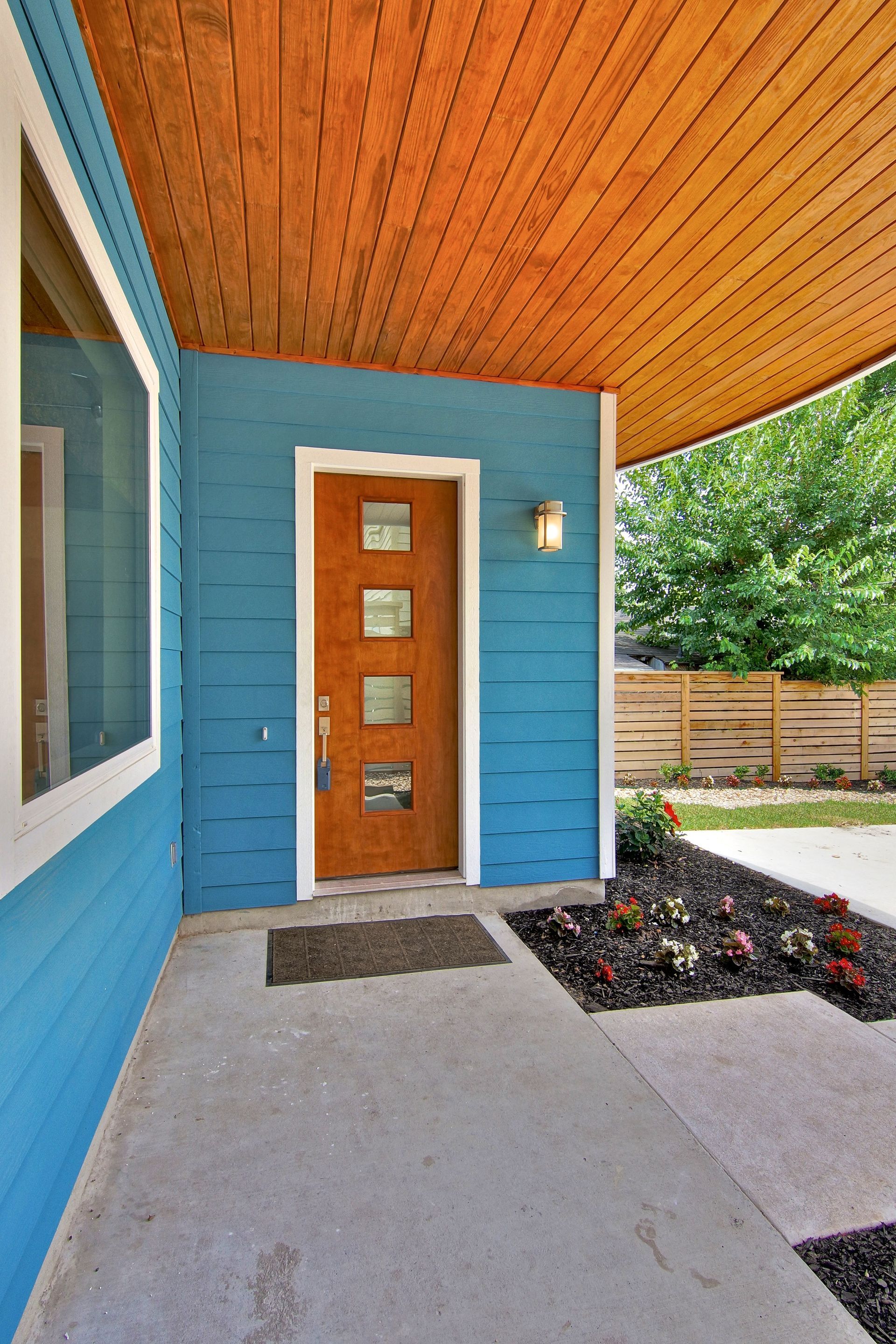 A bright blue home entrance with a wood-paneled ceiling, a modern wooden door with four glass insets, and a concrete porch.