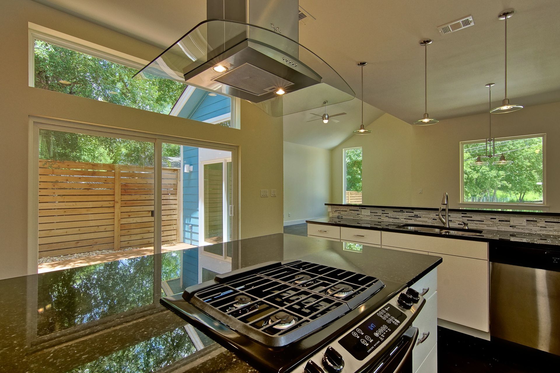 Modern kitchen with a dark granite island cooktop, stainless steel range hood, white cabinets, and large windows.