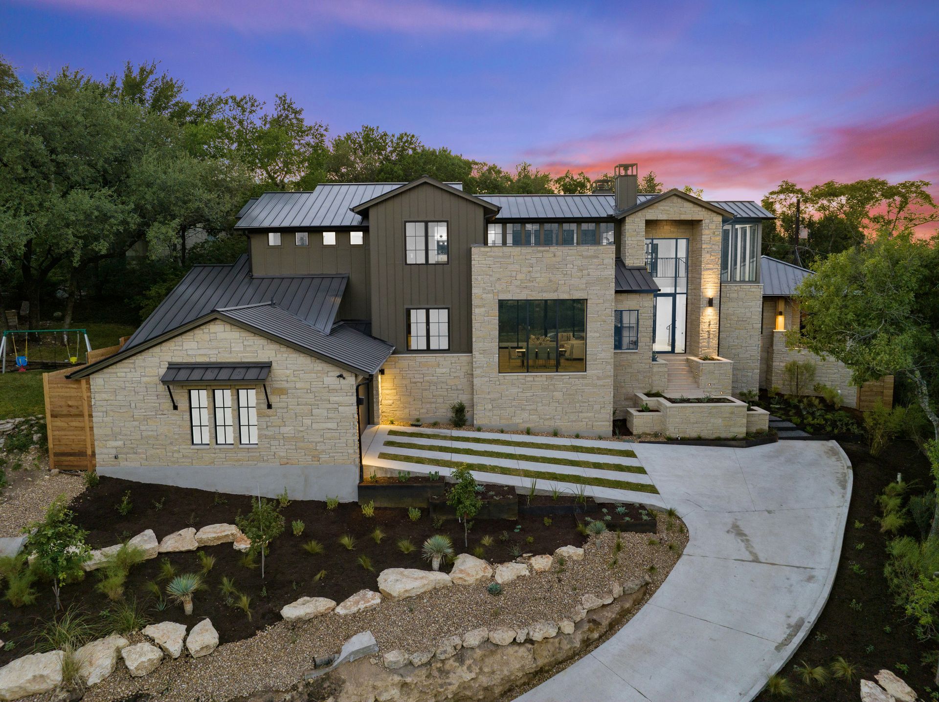 A modern, two-story home with stone and dark metal siding, featuring a metal roof, curved driveway, and sunset backdrop.