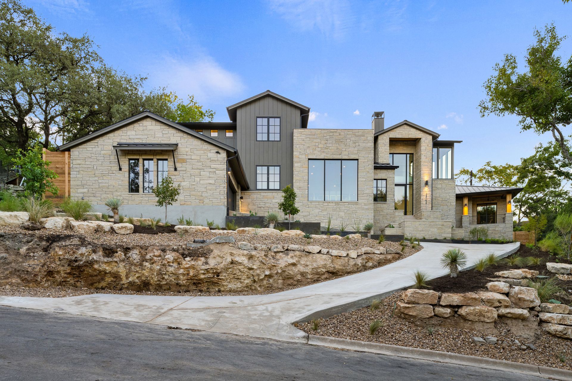 A modern, multi-level stone house with a concrete driveway, surrounded by trees and tiered landscaping on a sunny day.