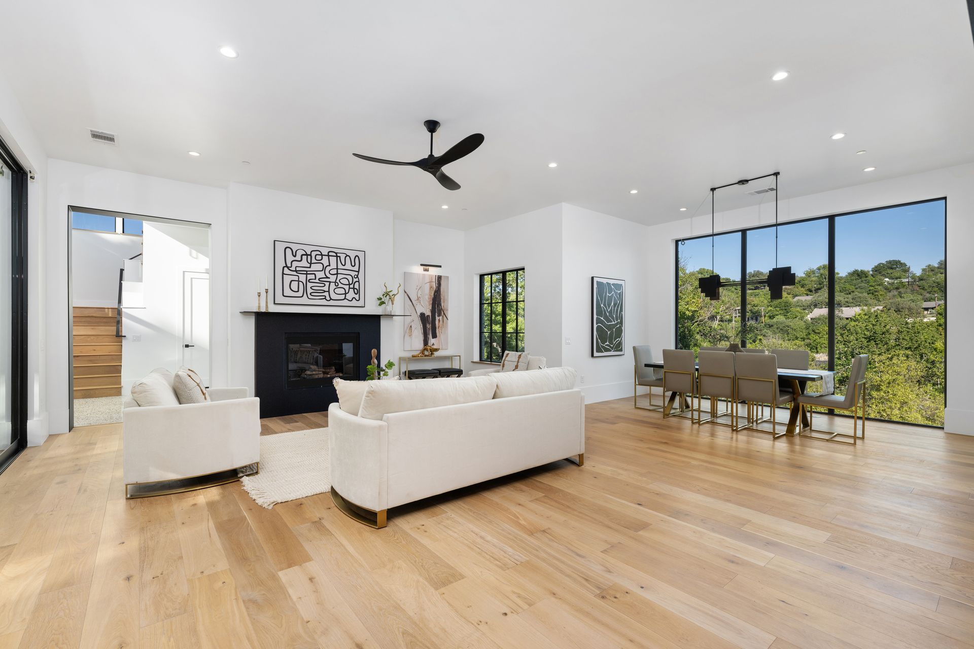 A modern living and dining room with white furniture, a black fireplace, hardwood floors, and large windows with greenery.