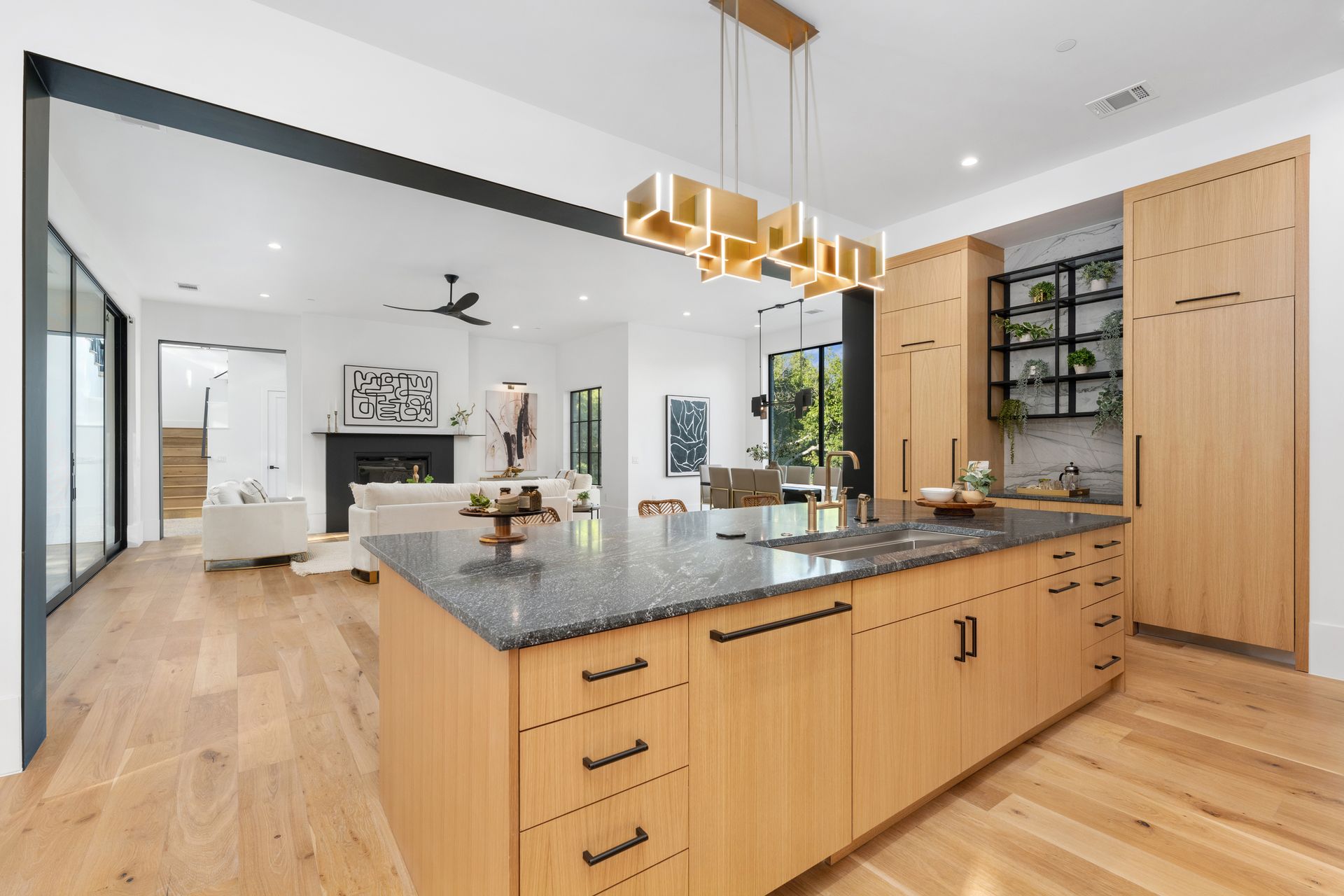 Modern kitchen with light wood cabinets, granite island, and gold chandelier, opening into a living room with a fireplace.