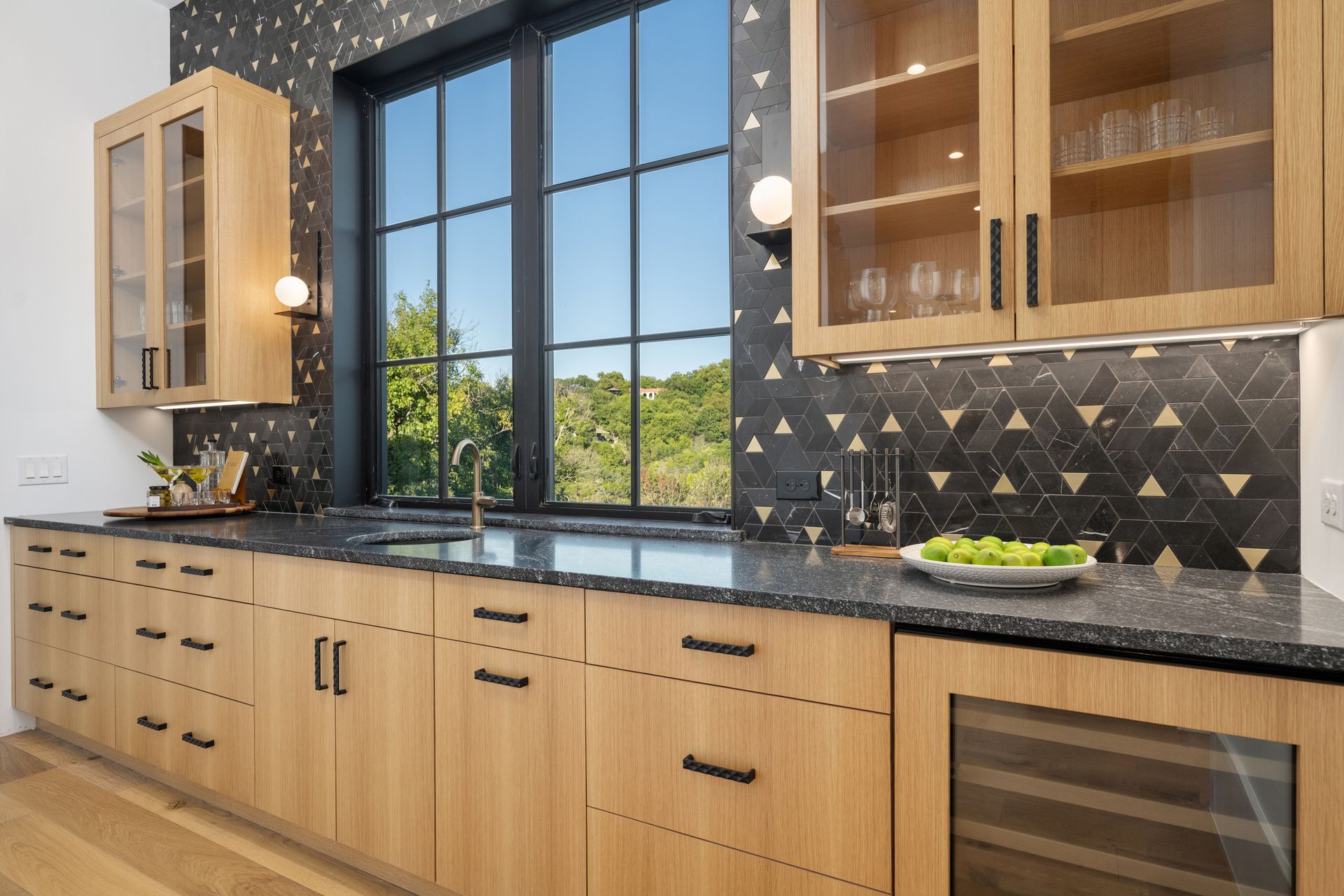 Modern kitchen with light wood cabinets, dark granite countertops, black geometric backsplash, and a window view.