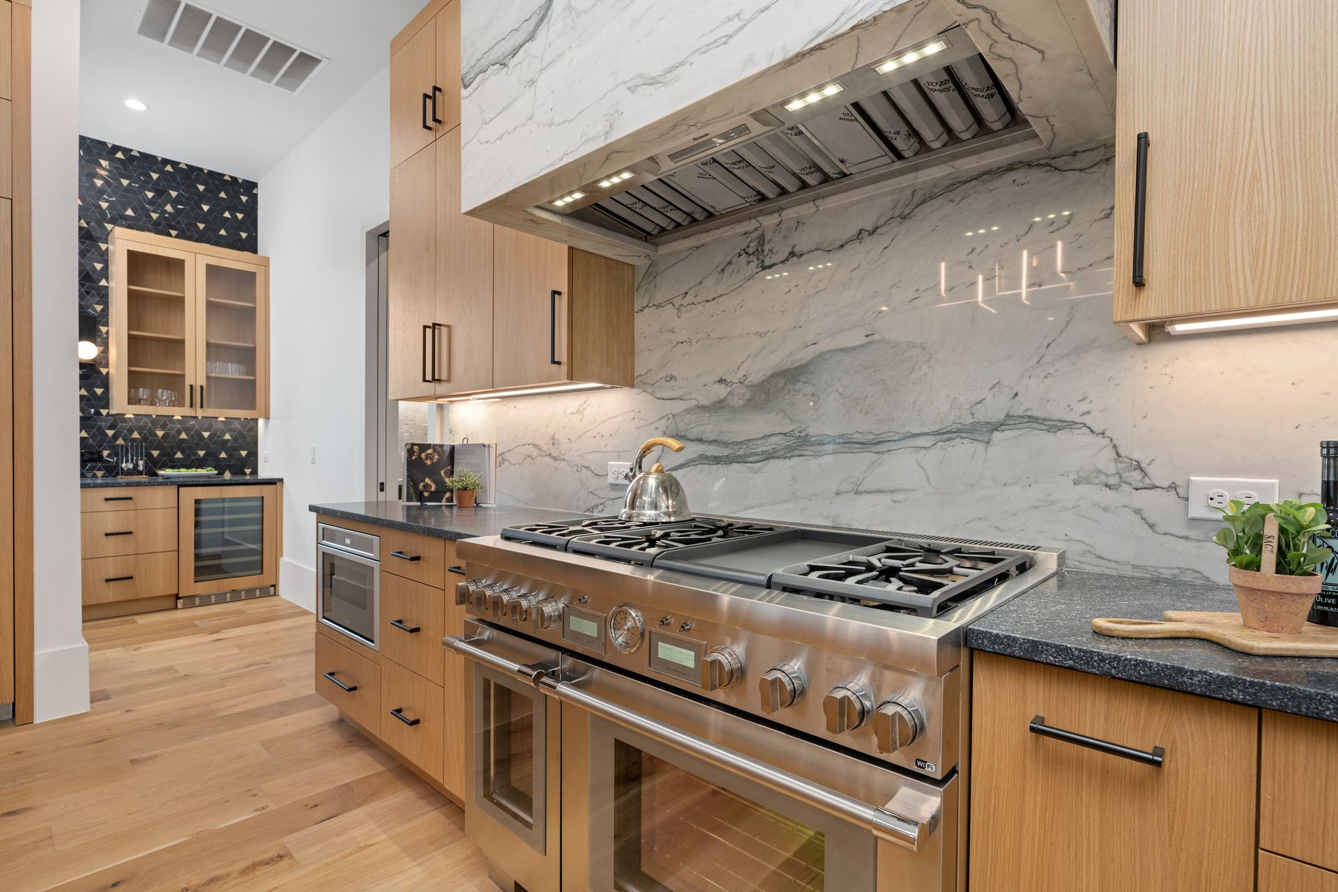 A modern kitchen featuring light wood cabinets, marble backsplash, a stainless steel gas range, and a separate wet bar.