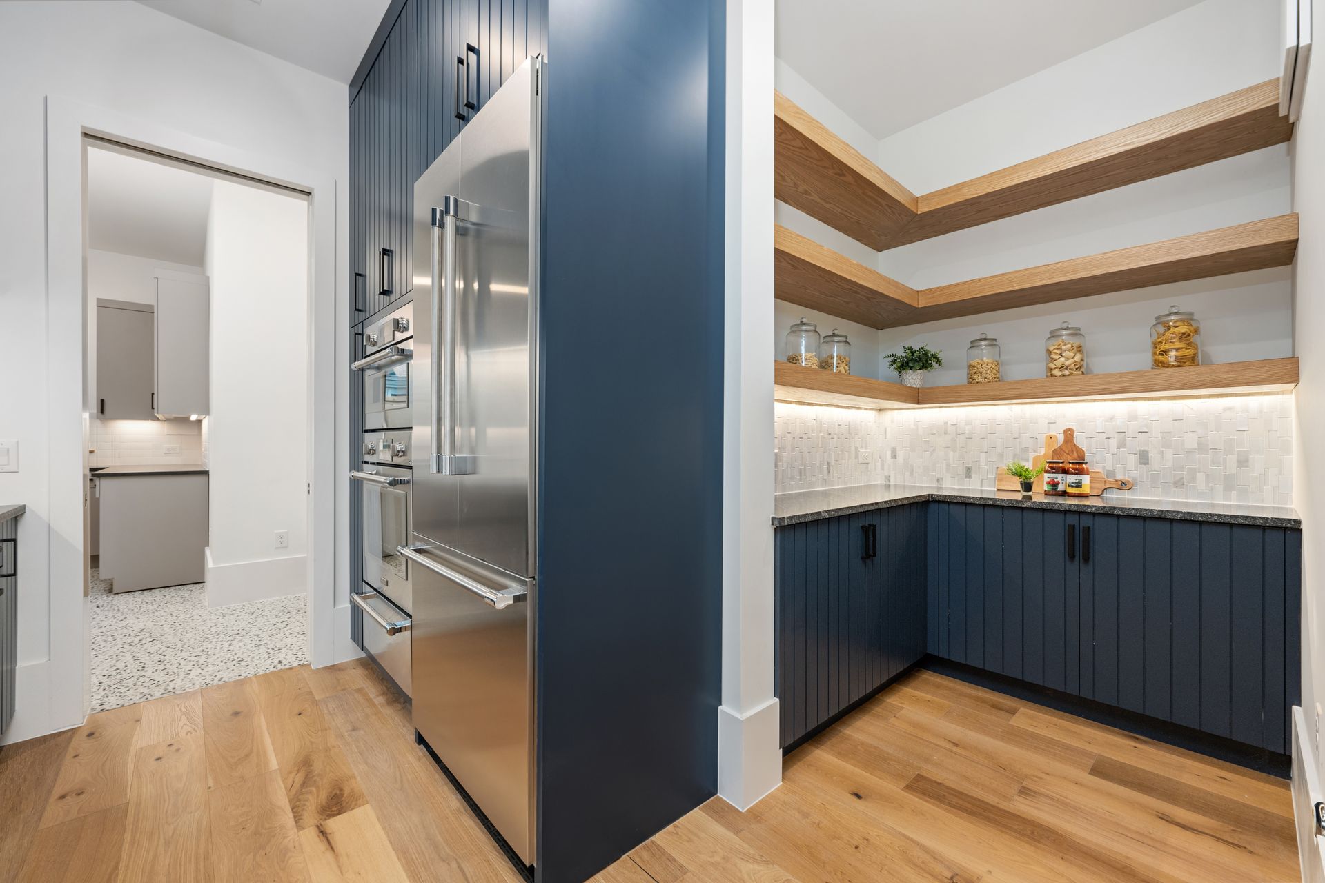 A modern pantry with navy blue cabinets, light wood flooring, and shelves, next to a stainless steel refrigerator.