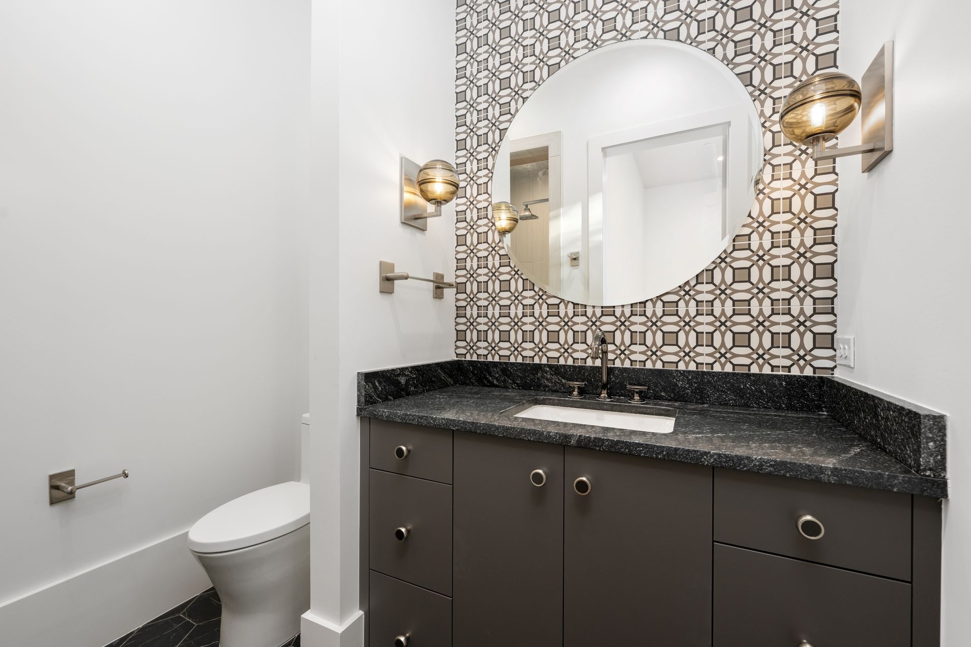 A modern powder room with a dark grey vanity, black speckled countertop, round mirror, and a geometric patterned tile wall.