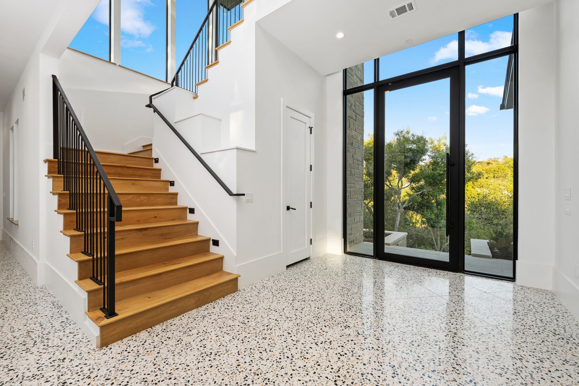 A modern foyer features a wooden staircase with a black railing, terrazzo flooring, and a large black-framed glass door.