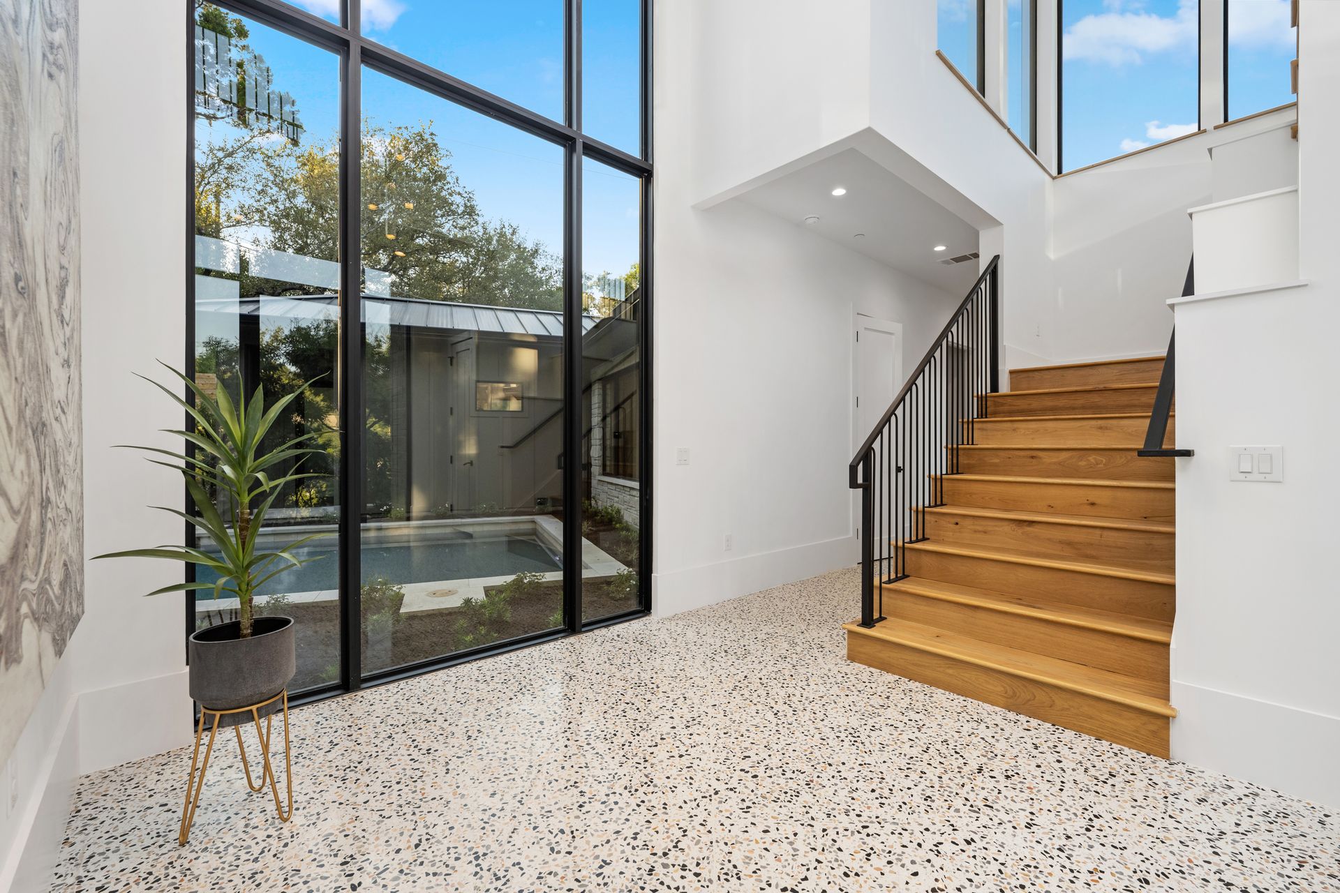 Modern entryway with high ceilings, tall windows overlooking a pool, terrazzo flooring, and wooden stairs with iron railing.