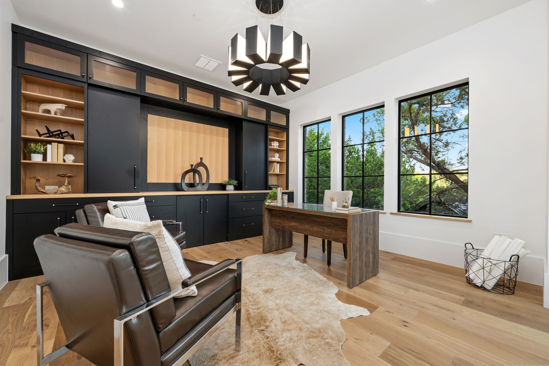 A home office featuring a desk, leather chairs, a cowhide rug, built-in black shelving, and large windows with garden views.