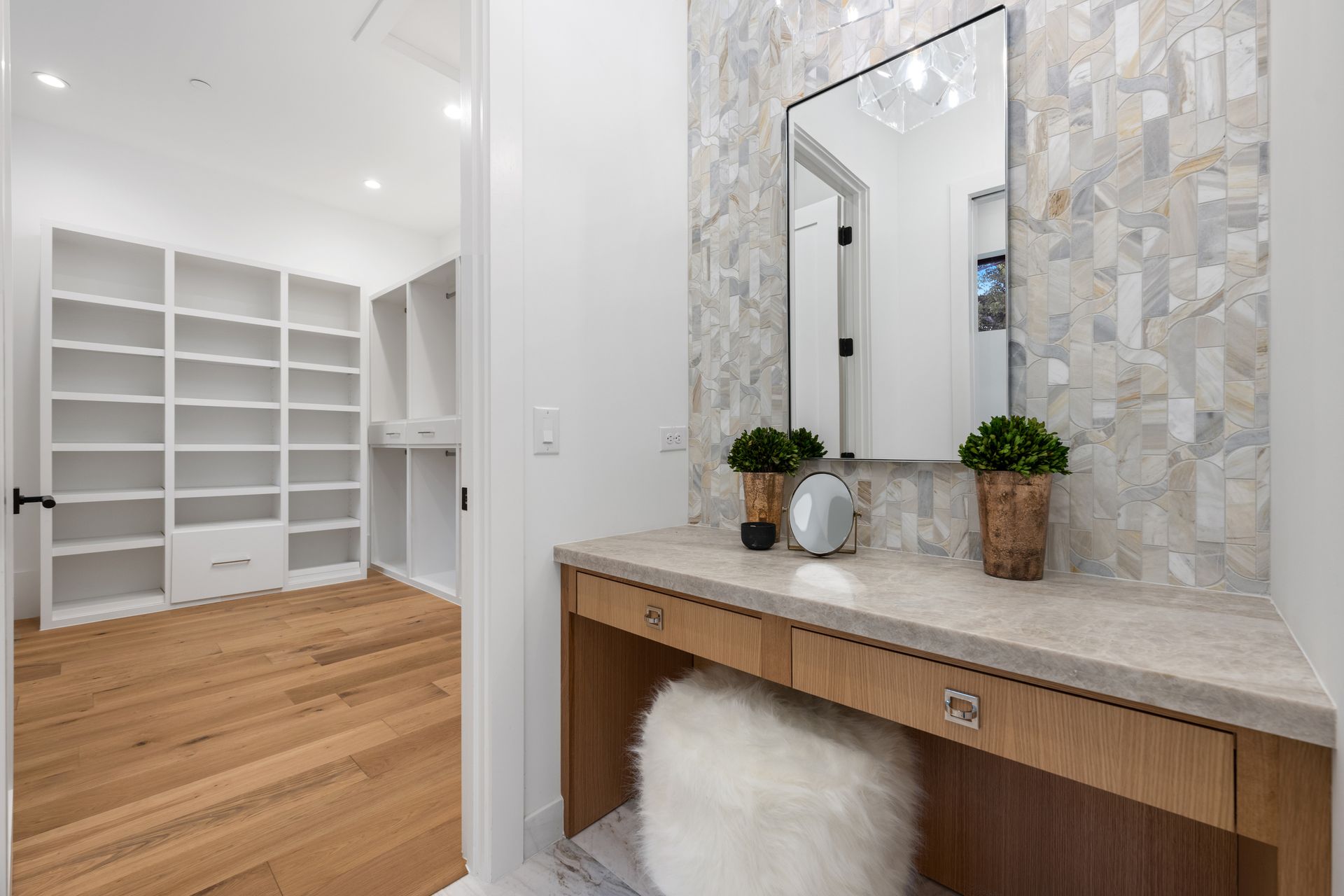 A vanity with a stone countertop and mirror, positioned beside a doorway leading to a brightly lit, empty walk-in closet.