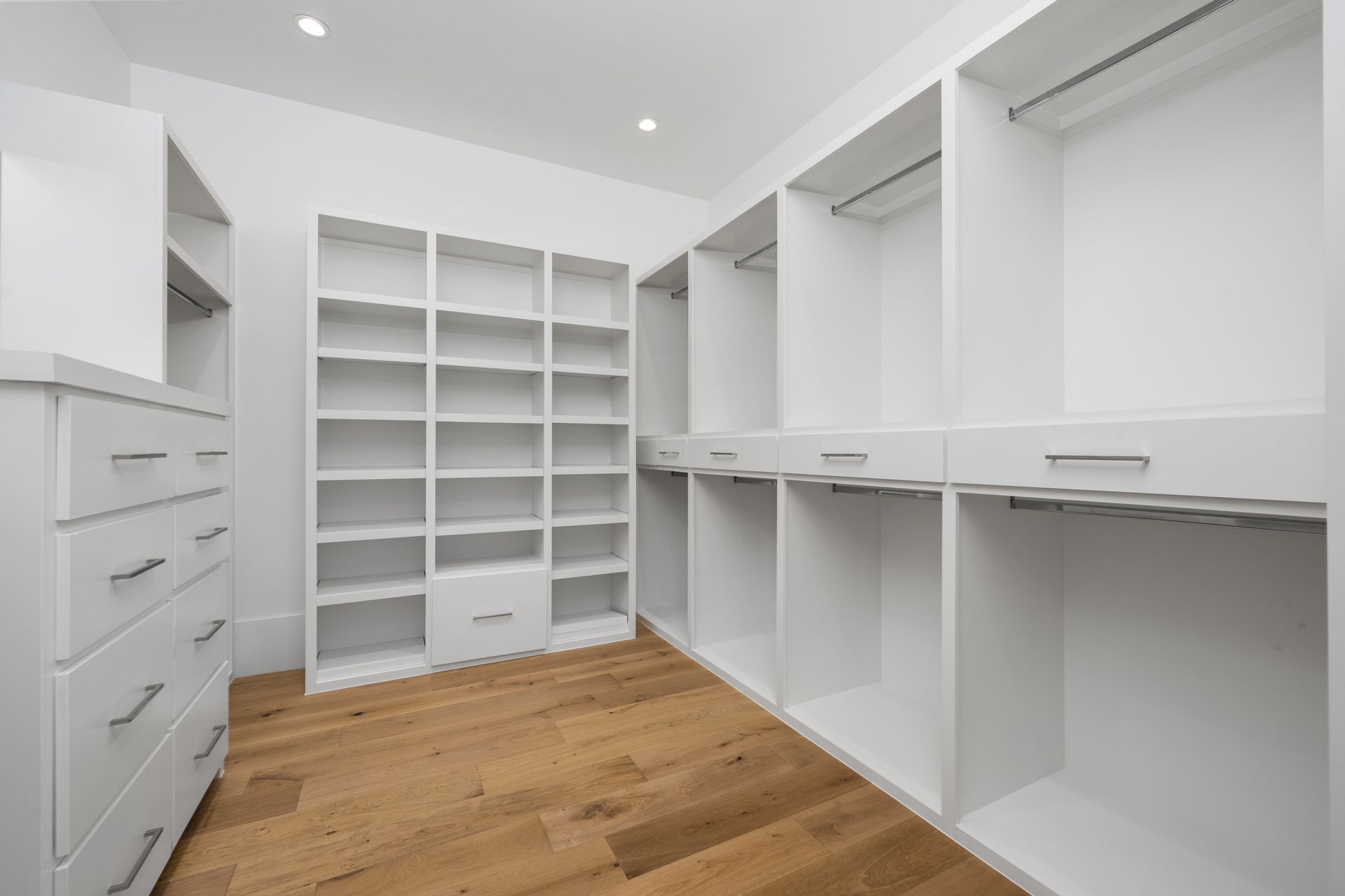Walk-in closet with white shelving, cabinets, and hanging rods over light wood flooring in a bright, modern room.