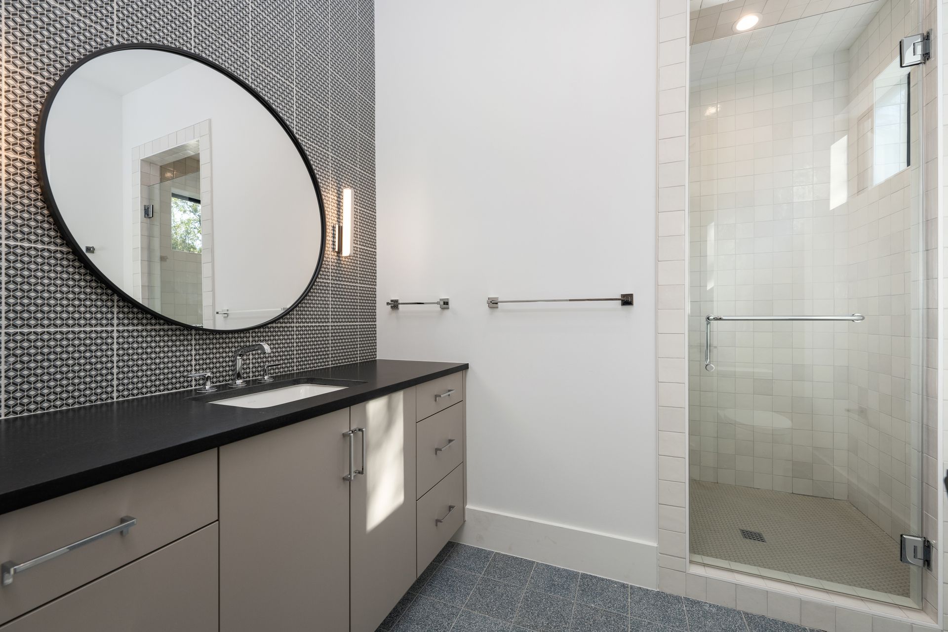 Modern bathroom with a tan vanity, black countertop, round mirror, patterned wall, and a glass-enclosed shower.