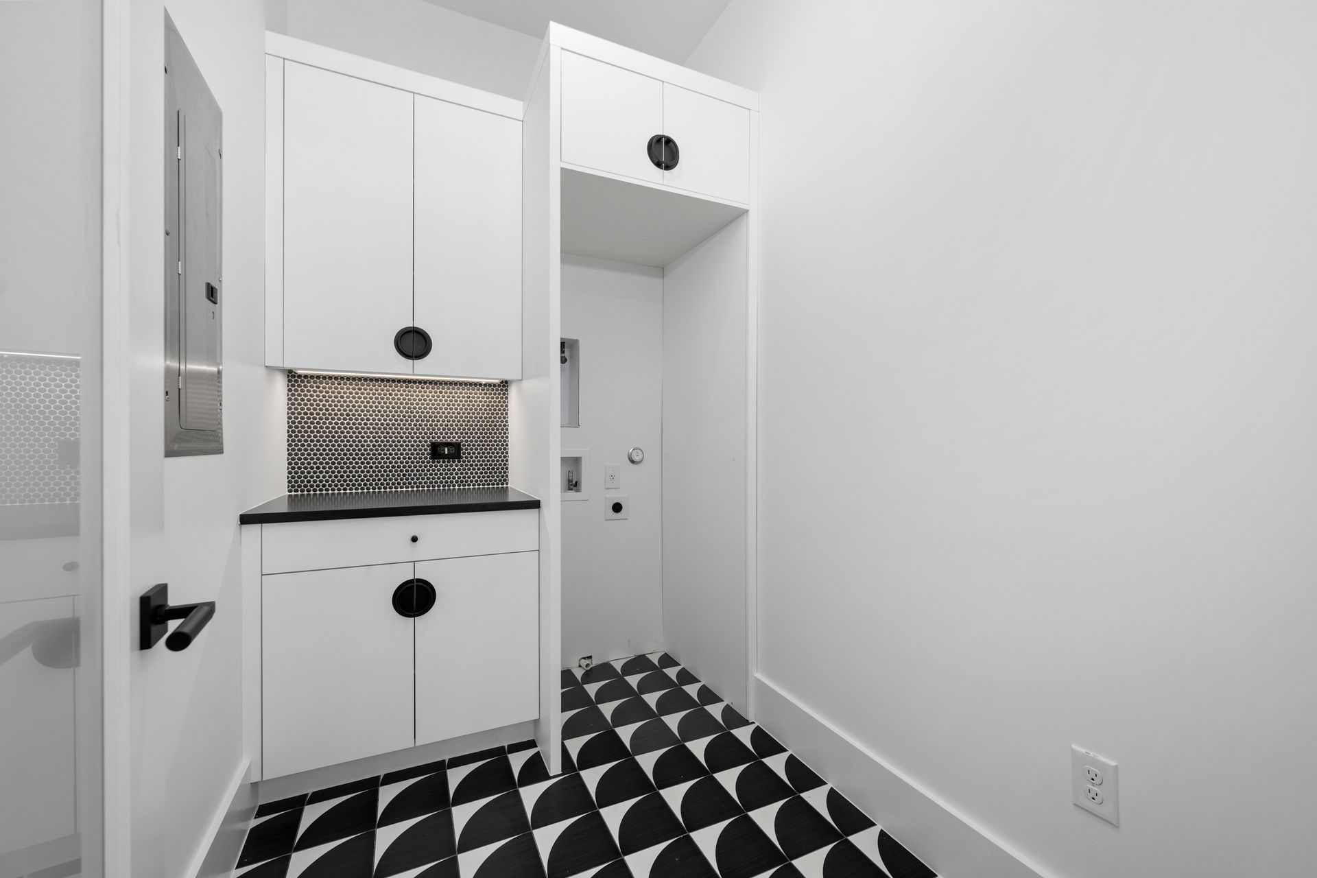 A modern white laundry nook with patterned black and white tile flooring, white cabinets, and a textured metal backsplash.