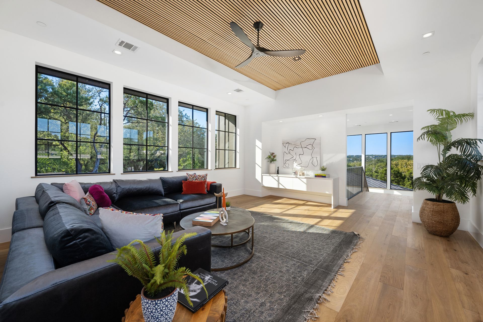 A modern living room with a black sectional sofa, a textured wood-patterned ceiling, and large windows with garden views.