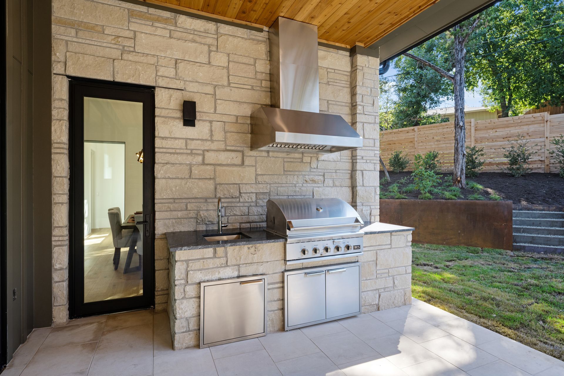 Outdoor kitchen with a stone-clad grill, stainless steel hood, and access door on a patio by a grassy backyard.