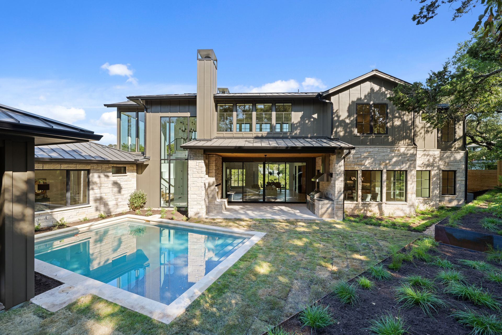 Modern two-story house with stone and dark trim, featuring a backyard pool and patio under a bright, sunny sky.