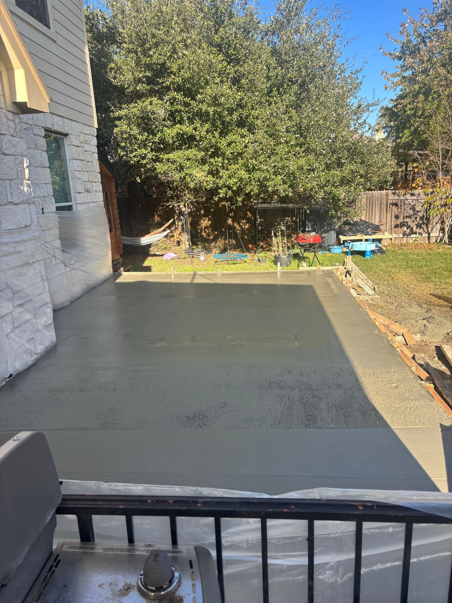 A newly poured, flat concrete patio extends from the side of a white stone house into a sunny backyard.