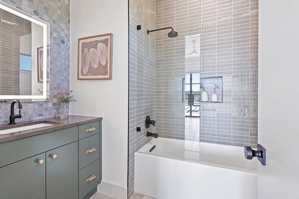 A bathroom with a sage green vanity, a lighted mirror, and a white bathtub with light gray tiled walls and black fixtures.