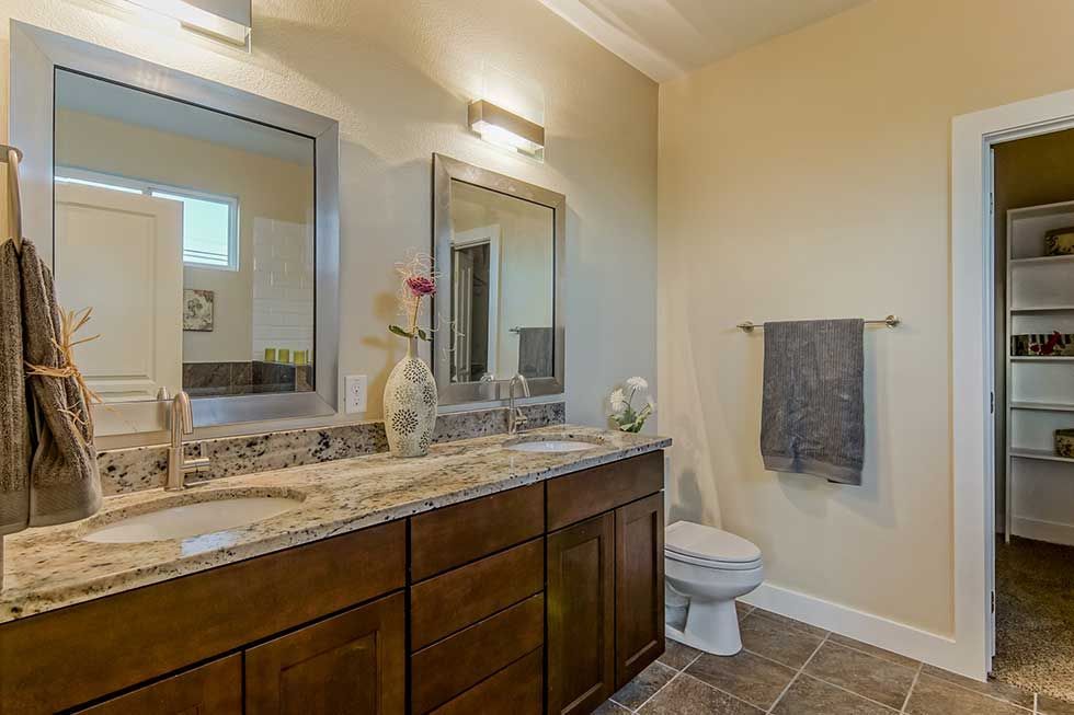 A bathroom vanity with dark wood cabinets, a speckled granite countertop, dual sinks, mirrors, and a toilet.