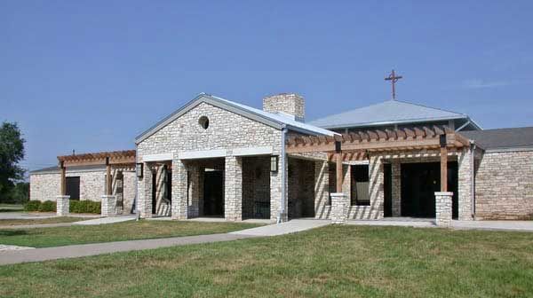 A stone church building under a clear blue sky, featuring a central gabled entryway and a cross atop the roof.