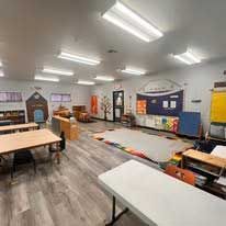 A classroom with light wood-look flooring, multiple tables, chairs, a rug in the center, and a large display board on a wall.
