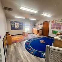 A brightly lit preschool classroom with a blue alphabet rug, a colorful area rug, and wood-toned storage furniture.