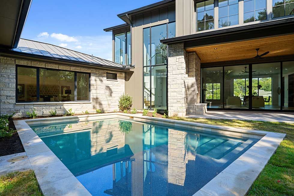 A modern house with stone walls and large windows overlooks a rectangular swimming pool on a sunny day.