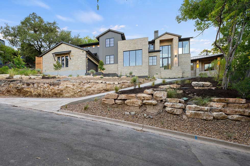 A two-story modern stone house with gray accents, glass windows, and a landscaped front yard with stone retaining walls.