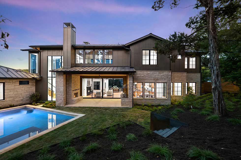 A two-story modern home at twilight with a pool, stone and dark metal siding, and a covered patio opening to the yard.