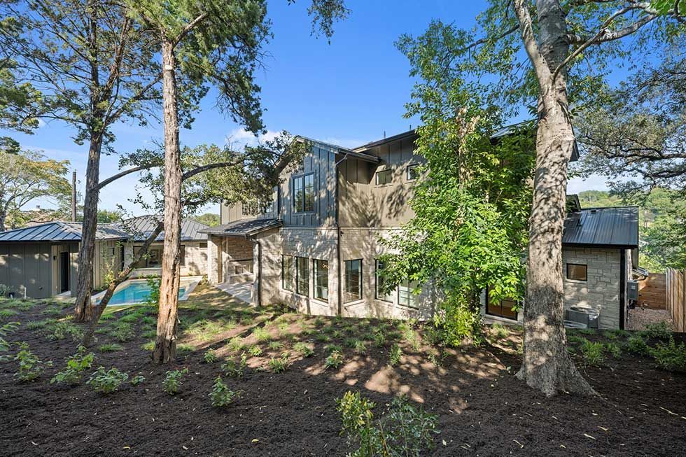 A two-story stone and dark metal house with a swimming pool, surrounded by trees on a sunny day.