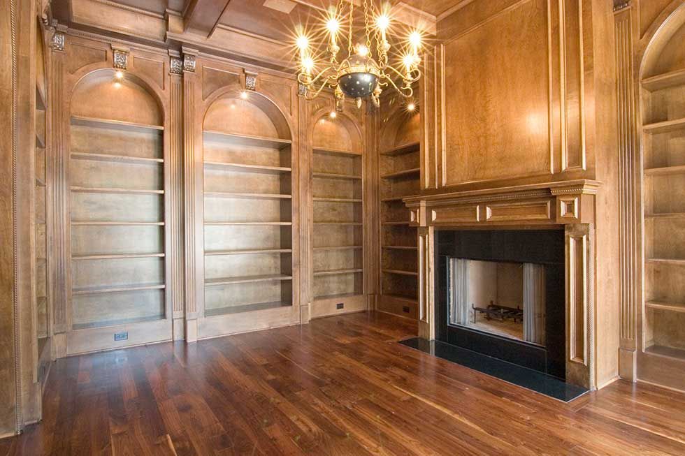 A library room with wood-paneled walls, built-in arched bookshelves, a black fireplace, and a hanging chandelier.