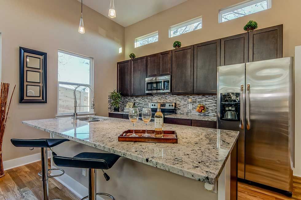 A modern kitchen with dark wooden cabinets, granite countertops, a stainless steel refrigerator, and two bar stools.
