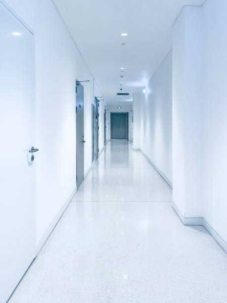 A long, bright white, sterile hospital hallway with polished floors and several closed doors on the left side.