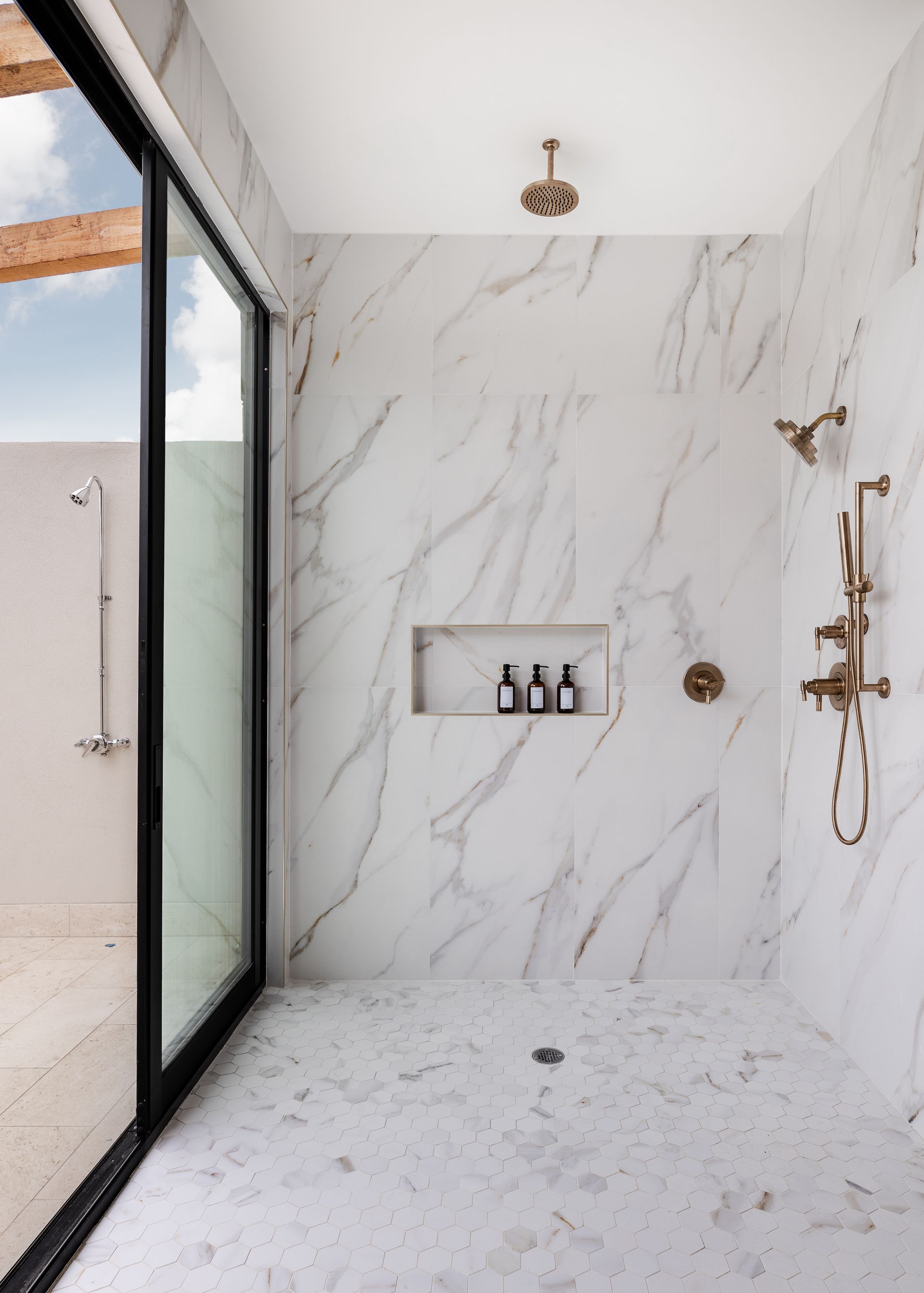 A modern walk-in shower with white marble walls, gold fixtures, a recessed shelf, and a large sliding glass door.