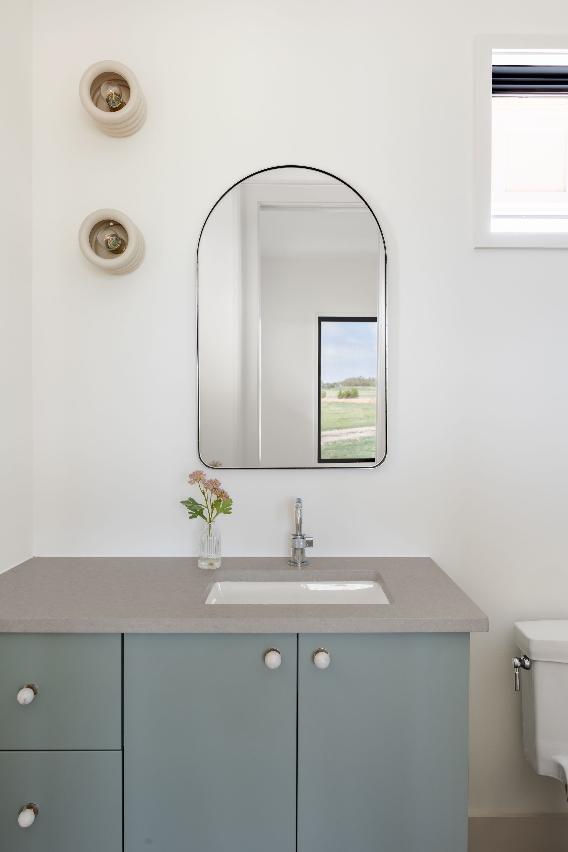 A bathroom vanity with a gray-blue cabinet, a gray countertop, a rectangular sink, and an arched wall mirror.