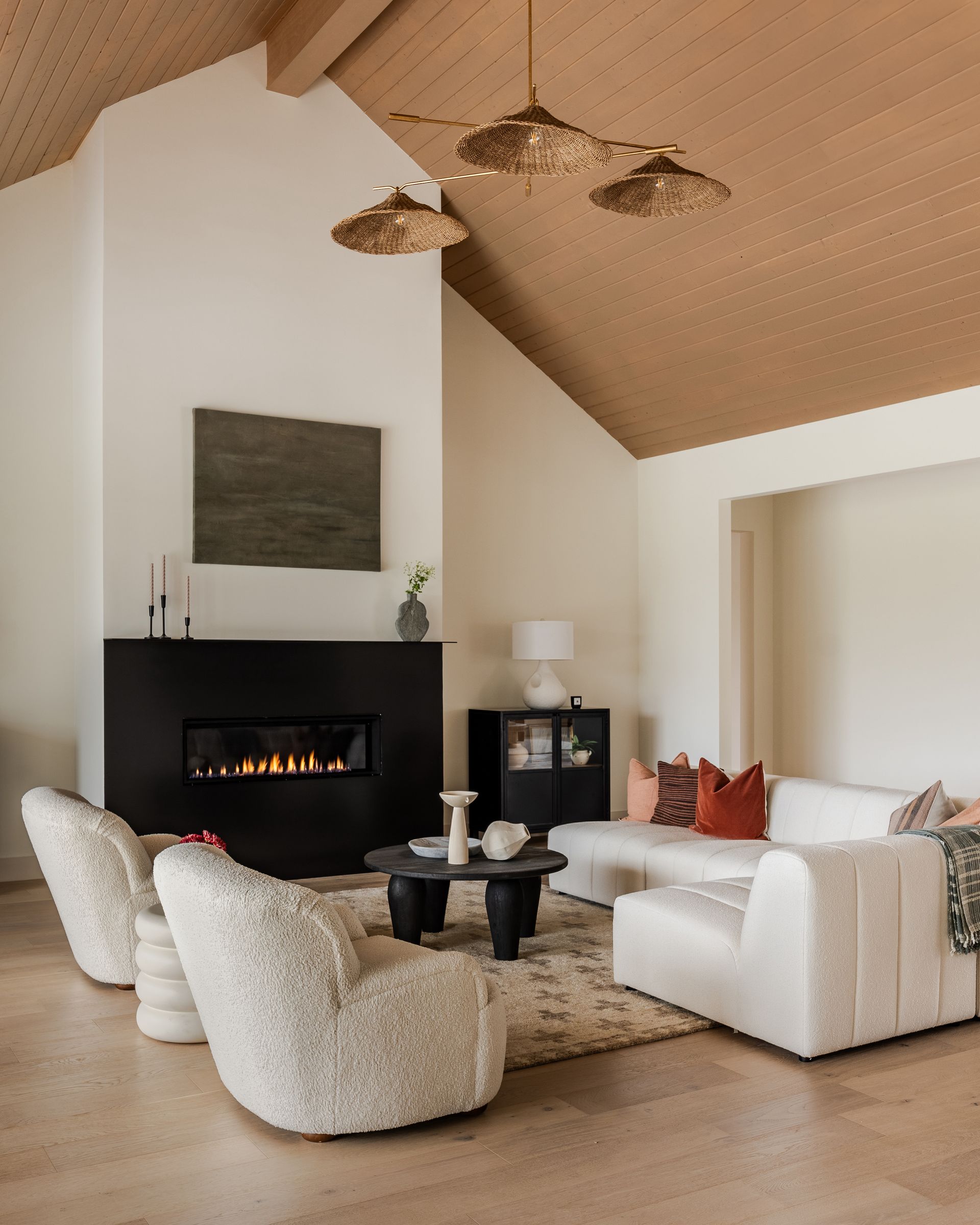 A modern living room with a black fireplace, textured cream chairs, a white sofa, and a wood-paneled vaulted ceiling.