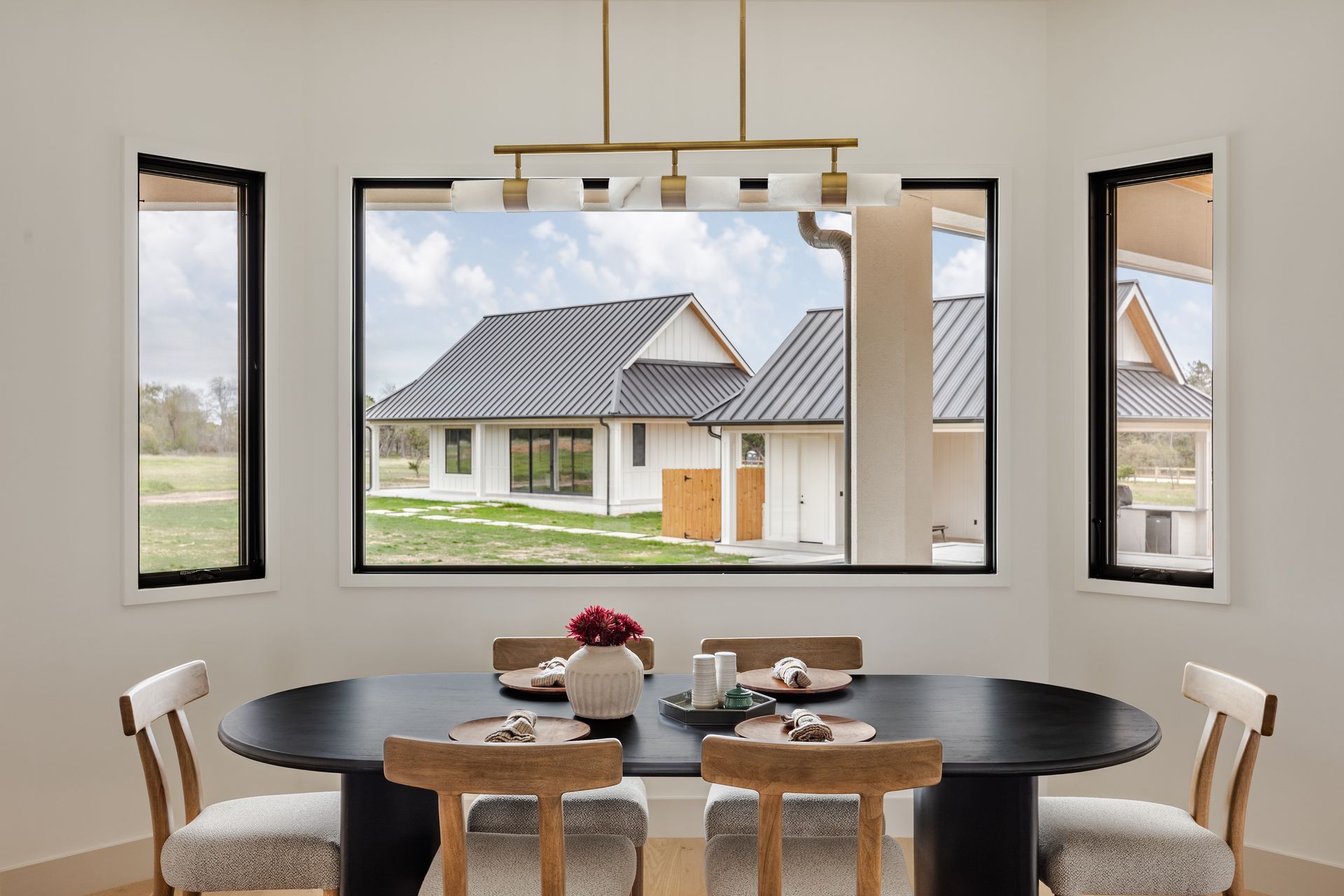 A dining room with a dark oval table, four light wood chairs, and a modern light fixture overlooking a rural house.