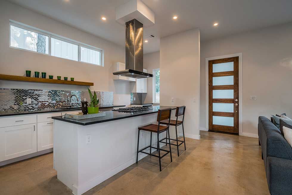 Modern kitchen with a white island, stainless steel range hood, wooden shelving, and a glass-paneled wooden door.
