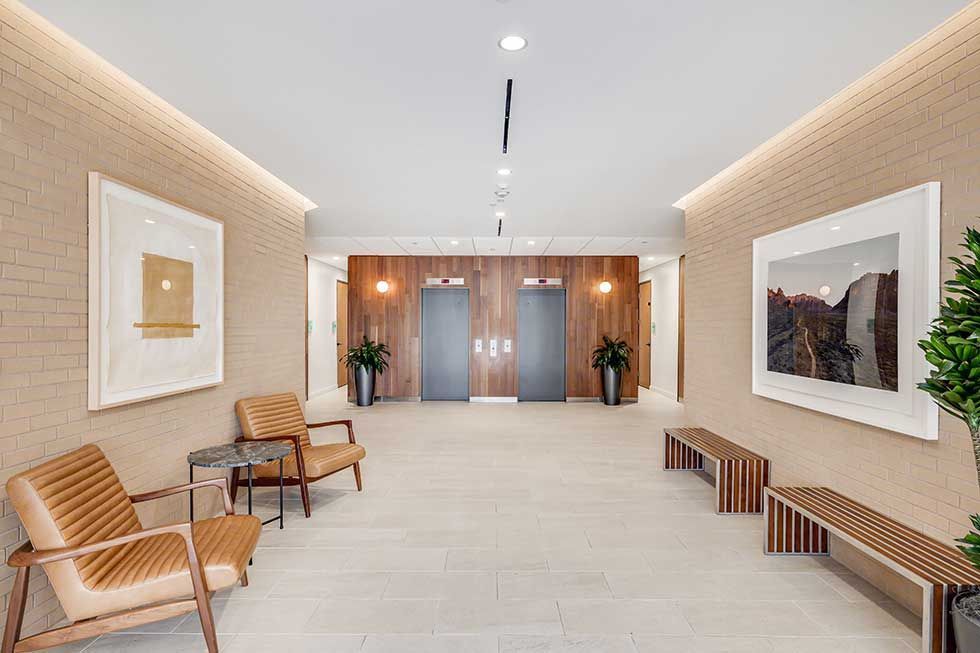 A modern, minimalist lobby featuring two elevators, tan brick walls, two armchairs, benches, and framed wall art.